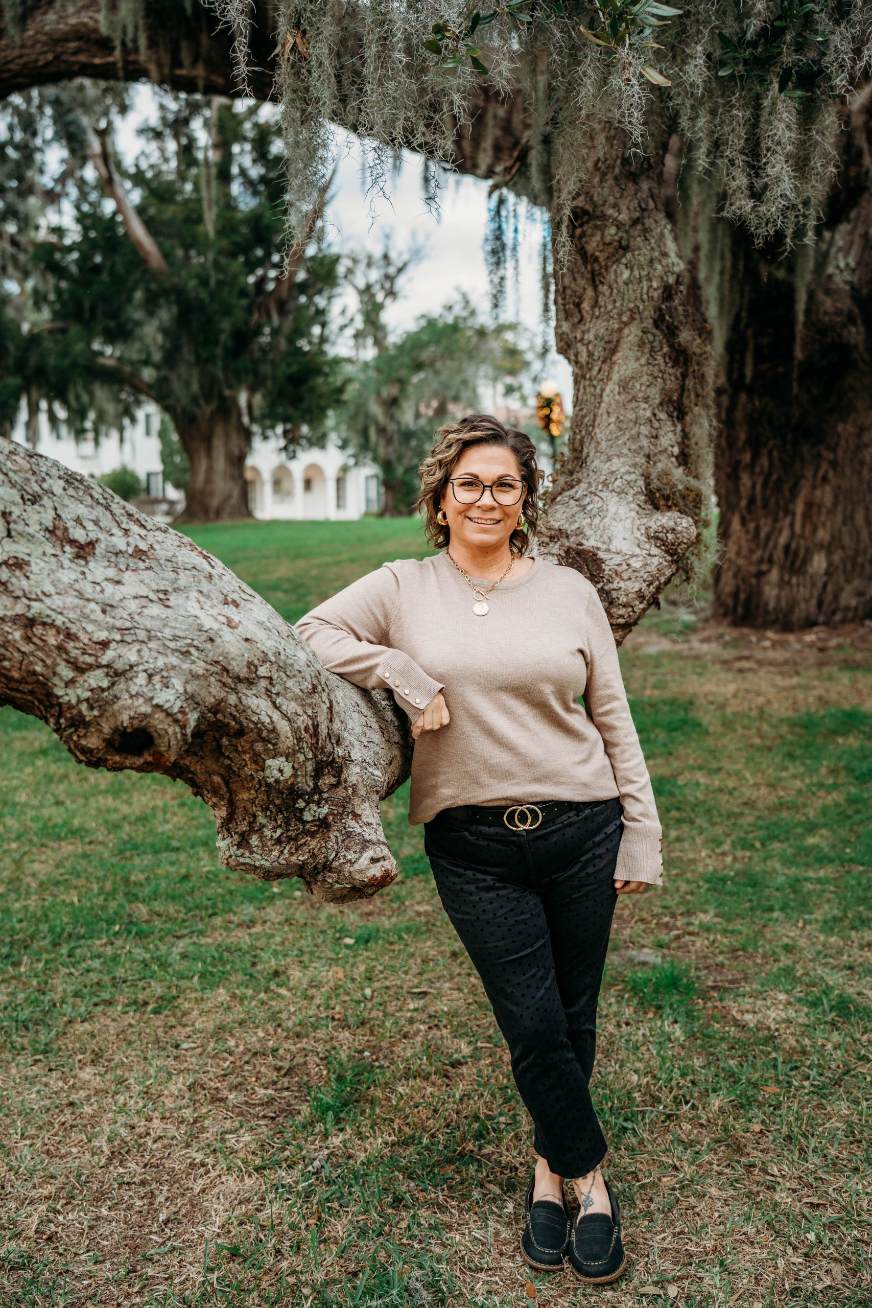 Woman in a tan top and dark pants leaning on a life oak branch.